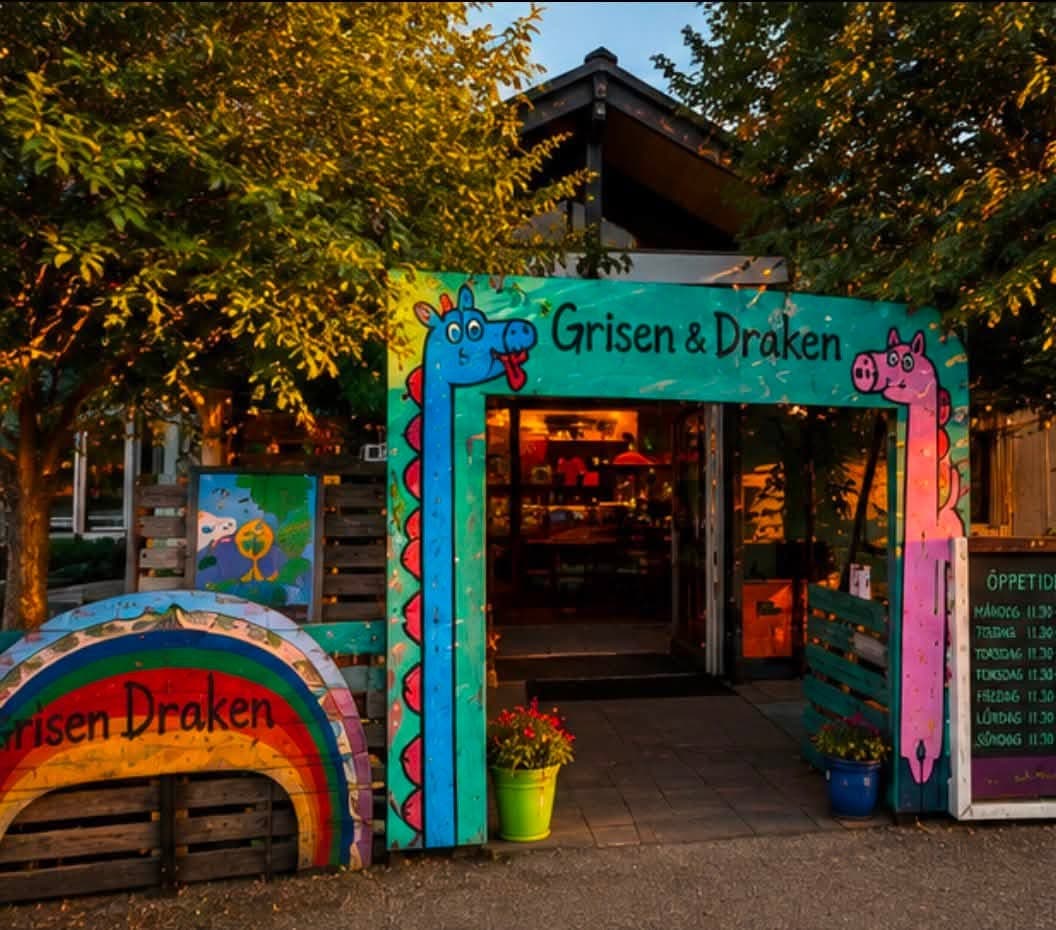 Entrén till Grisen & Draken i Brösarp en sommarkväll: en handmålad portal där draken och grisen ramar in dörren, med regnbågsskylten till vänster och öppettidstavlan till höger.