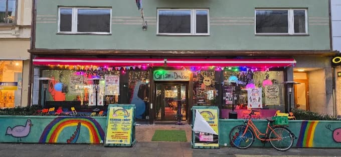 Entrén till Grisen & Draken på Östra Boulevarden i Kristianstad: skyltfönster fulla av affischer och ljusslingor under en röd markis, regnbågar, en gris och en drake målade på muren utanför, och en cykel parkerad vid dörren.